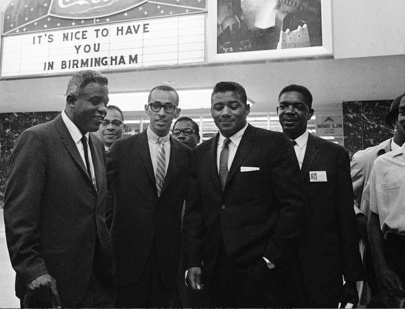 Athletes Jackie Robinson (left) and Floyd Patterson (third from left) are welcomed to riot-torn Birmingham, Ala., on May 13, 1963, by Wyatt Tee Walker (second from left) and a sign overhead. They came here to address an integration rally. (AP Images)